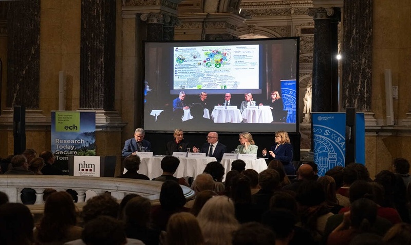Eine Podiumsdiskussion mit sechs Personen, die an einem Tisch auf einer Bühne sitzen, während ein großes Publikum zuschaut; im Hintergrund ist eine Leinwand mit einer Präsentation zu sehen.