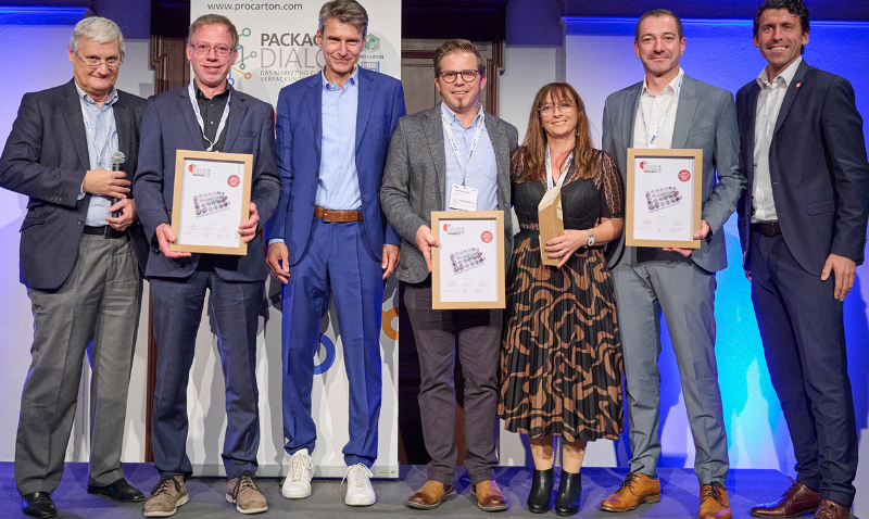 Eine Gruppe von sieben Personen steht auf einer Bühne. Drei von ihnen halten gerahmte Urkunden in den Händen. Alle sind formell gekleidet und lächeln. Der Hintergrund ist blau beleuchtet.