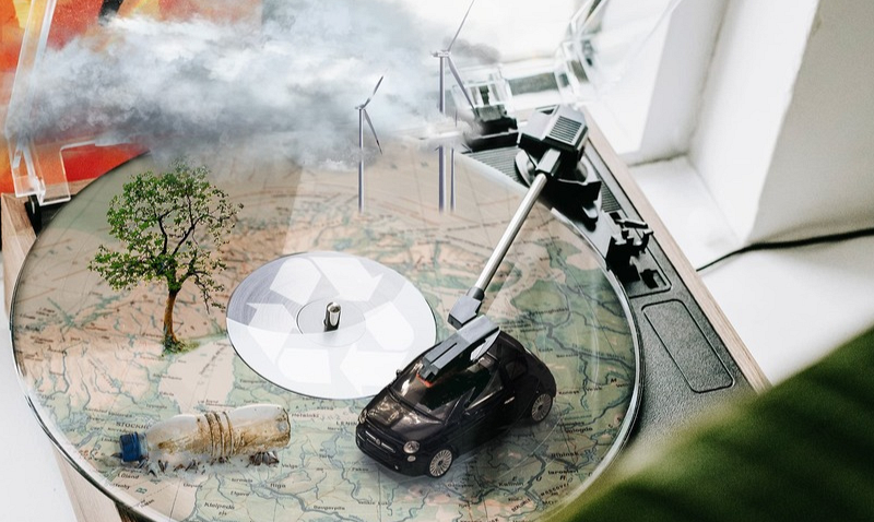 Ein Plattenspieler mit einer kreativen Szene darauf: ein kleines Auto, ein Baum, Windräder und eine Plastikflasche auf einer Landkartenoberfläche, umgeben von künstlichen Wolken.